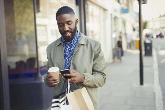 Young man with coffee and shopping bags texting with cell phone on urban sidewalk