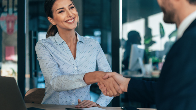 A businesswoman and a businessman sitting at a table shaking hands.