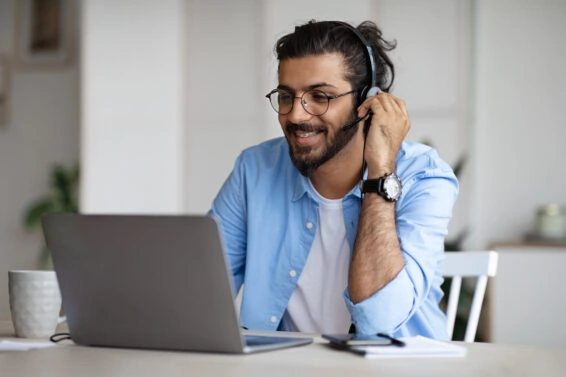 Smiling young indian guy watching webinar on laptop, wearing headset and eyeglasses